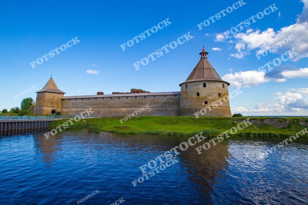 На фотографии изображена крепость Орешек, расположенная на острове в истоке реки Невы, близ Ладожского озера. Массивные стены и башни отражают богатую историю этого оборонительного сооружения. Голубое небо и зелёная растительность подчёркивают красоту окружающего пейзажа. Крепость является важным памятником истории и архитектуры, привлекающим туристов и любителей истории.